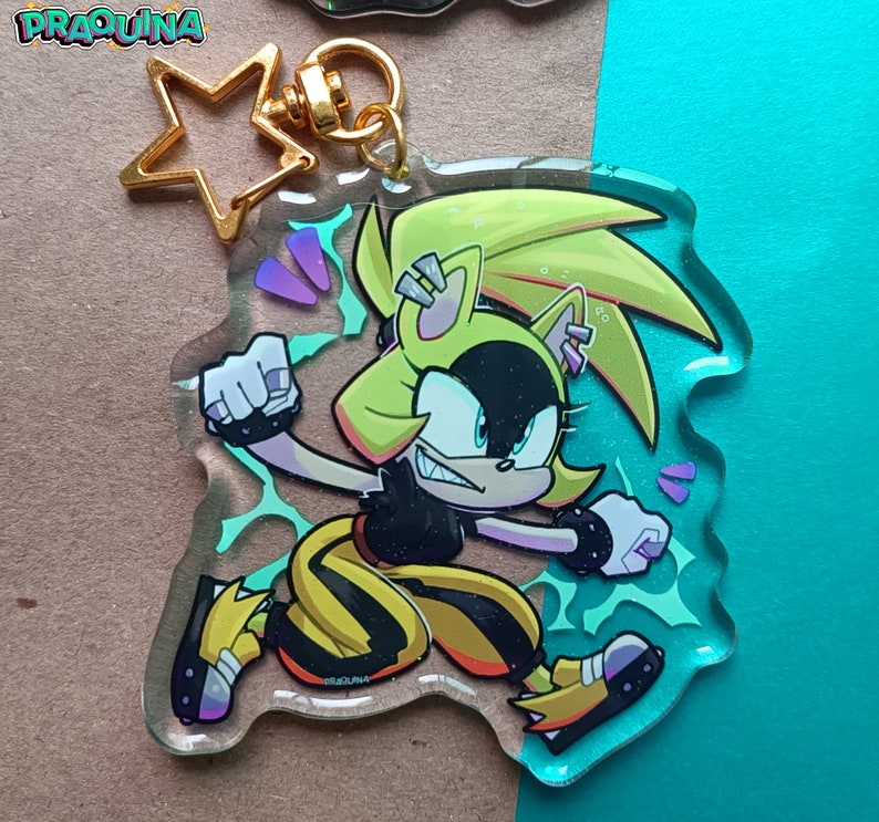Op de afbeelding: Een geel en zwart cartoonfiguur met stekelig geel haar en een zwart masker. Het figuur staat in een vechtpose met een geel en zwart pak. Het figuur zit op een transparante acryl sleutelhanger met een gouden sterbedel.