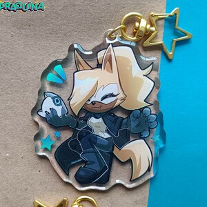Op de afbeelding: Een transparante acryl sleutelhanger met een cartoonfiguur met blond haar, die een zwart jack draagt en een klein wit voorwerp vasthoudt. Het figuur staat op een blauwe en witte achtergrond met sterren.
