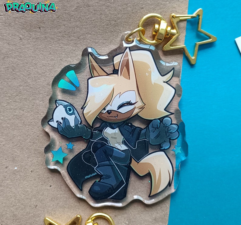 Op de afbeelding: Een transparante acryl sleutelhanger met een cartoonfiguur met blond haar, die een zwart jack draagt en een klein wit voorwerp vasthoudt. Het figuur staat op een blauwe en witte achtergrond met sterren.