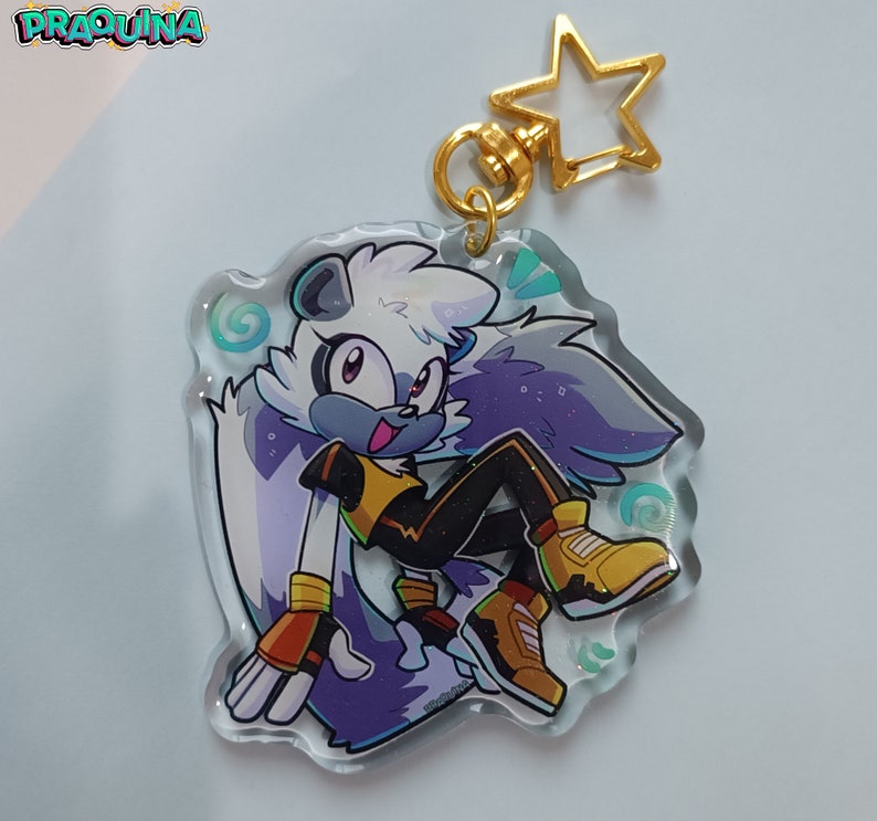 Op de afbeelding: Een transparante sleutelhanger met een gouden sterbedel waarop een paars-wit cartoonfiguur met een geel-zwarte outfit te zien is. Het figuur heeft een witte staart en draagt sneakers.