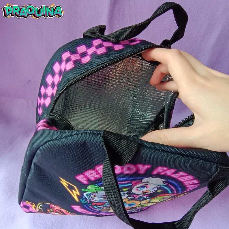 Peut inclure: Sac &agrave; d&eacute;jeuner noir avec un motif &agrave; carreaux rose et noir sur le dessus. Le sac a une fermeture &eacute;clair et une doublure isolante argent&eacute;e. Le devant du sac pr&eacute;sente un graphique de personnages du jeu vid&eacute;o "Five Nights at Freddy's" avec le texte "Freddy Fazbear's Mega Pizzaplex".