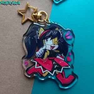 Op de afbeelding: Een transparante acryl sleutelhanger met een cartoonfiguur met zwart haar, gele vacht en een rood-zwarte outfit. Het personage knippert met zijn oog en wijst met &eacute;&eacute;n hand. De sleutelhanger heeft een gouden stervormige hanger bevestigd aan een metalen ring.