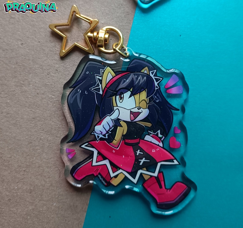 Op de afbeelding: Een transparante acryl sleutelhanger met een cartoonfiguur met zwart haar, gele vacht en een rood-zwarte outfit. Het personage knippert met zijn oog en wijst met &eacute;&eacute;n hand. De sleutelhanger heeft een gouden stervormige hanger bevestigd aan een metalen ring.