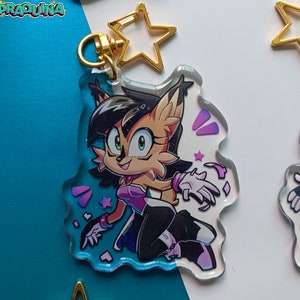Op de afbeelding: Een transparante acryl sleutelhanger met een cartoonfiguur met bruine vacht, zwart haar en groene ogen. Het personage draagt een paarse en roze outfit en is omringd door roze en paarse glitter. De sleutelhanger heeft een gouden sterbedel.