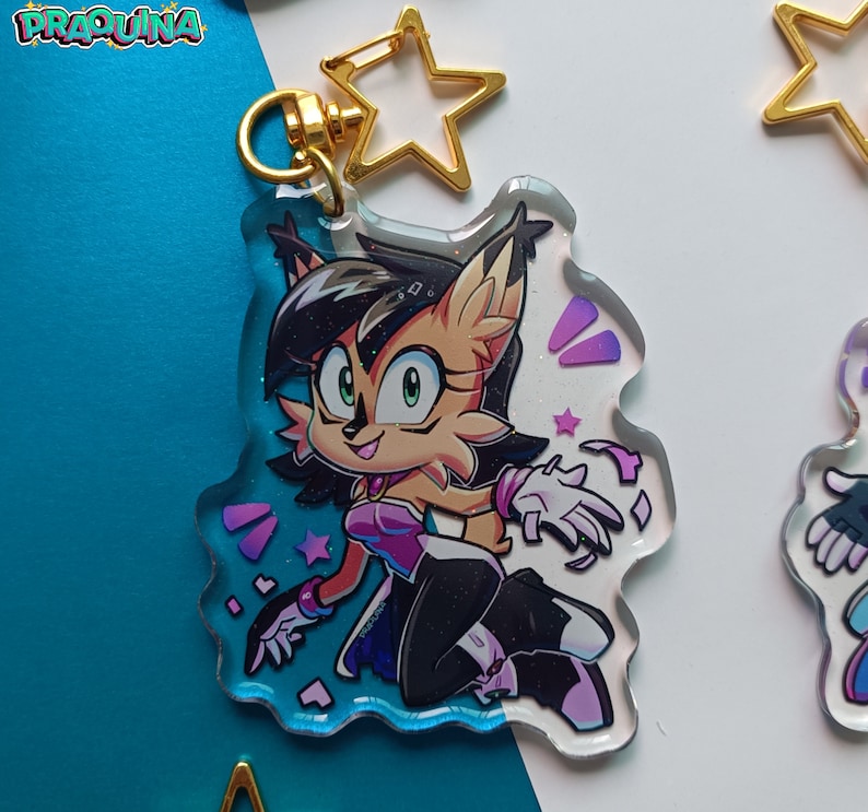 Op de afbeelding: Een transparante acryl sleutelhanger met een cartoonfiguur met bruine vacht, zwart haar en groene ogen. Het personage draagt een paarse en roze outfit en is omringd door roze en paarse glitter. De sleutelhanger heeft een gouden sterbedel.
