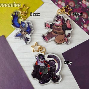 Op de afbeelding: Drie acryl sleutelhangers met stripfiguren. De sleutelhangers bevatten een blauwe vogel karakter met de naam "Doom", een varkens karakter met de naam "Pigtail" en een kat karakter met de naam "Kitty". Elke sleutelhanger heeft een gouden stervormige sluiting en de merknaam "PRAQUINA".