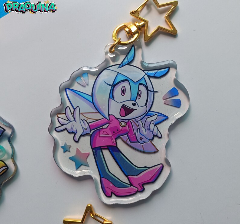 Op de afbeelding: Een transparante acryl sleutelhanger met een cartoonfiguur met blauwe ogen, witte vacht en roze kleding. Het personage heeft vleugels en houdt een pose aan met zijn armen uitgestrekt. De sleutelhanger heeft een gouden sterbedel.