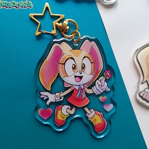 Op de afbeelding: Een transparante acryl sleutelhanger met een cartoonfiguur met cr&egrave;mekleurige vacht, roze oren en een rode jurk. Het personage houdt een roze bloem vast en heeft een gelukkige uitdrukking. De sleutelhanger heeft een gouden stervormige hanger en een metalen ring.