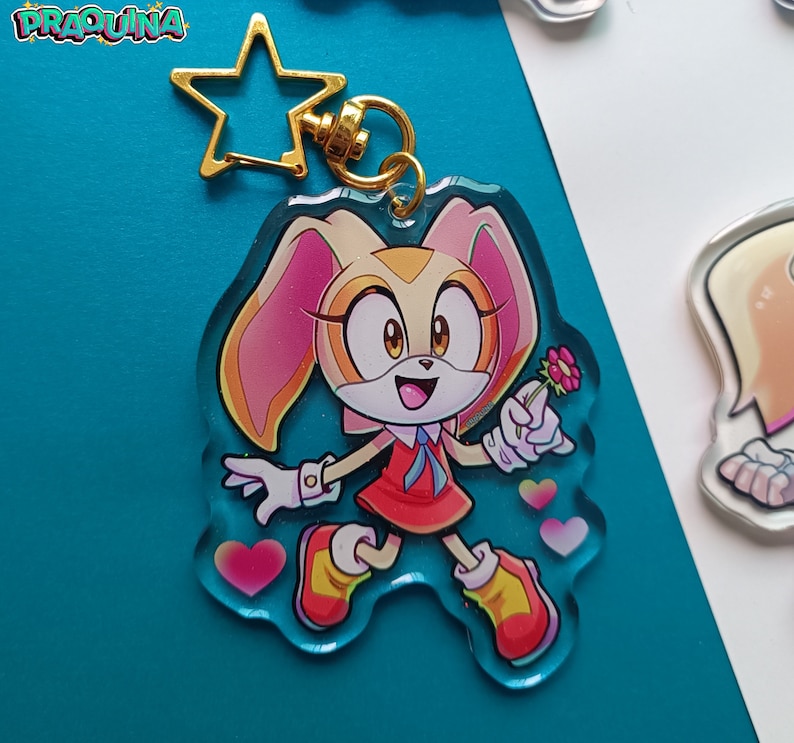 Op de afbeelding: Een transparante acryl sleutelhanger met een cartoonfiguur met cr&egrave;mekleurige vacht, roze oren en een rode jurk. Het personage houdt een roze bloem vast en heeft een gelukkige uitdrukking. De sleutelhanger heeft een gouden stervormige hanger en een metalen ring.
