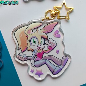 Op de afbeelding: Een wit en roze cartoon konijnenpersonage met groene ogen en een roze shirt. Het personage steekt zijn duim omhoog en heeft een glinsterende achtergrond. Het personage zit op een doorzichtige acryl sleutelhanger met een gouden ster hanger.