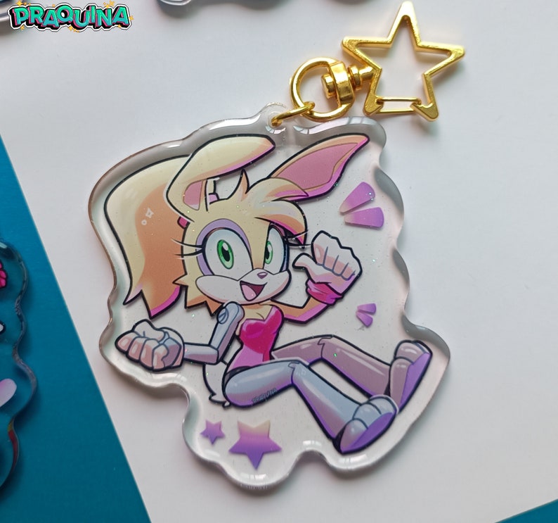 Op de afbeelding: Een wit en roze cartoon konijnenpersonage met groene ogen en een roze shirt. Het personage steekt zijn duim omhoog en heeft een glinsterende achtergrond. Het personage zit op een doorzichtige acryl sleutelhanger met een gouden ster hanger.