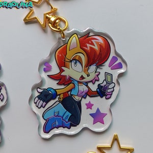 Op de afbeelding: Een transparante acryl sleutelhanger met een cartoonfiguur met rood haar, een blauw-witte outfit en een draagbare videogameconsole. Het personage is omringd door paarse en roze sterren op een witte achtergrond.