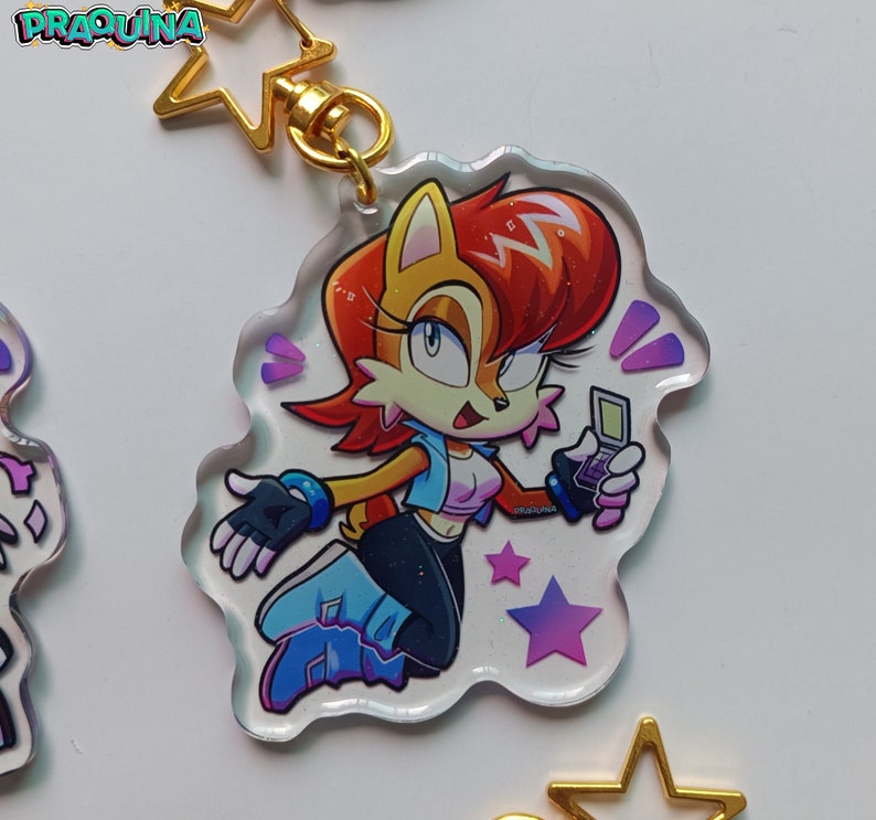 Op de afbeelding: Een transparante acryl sleutelhanger met een cartoonfiguur met rood haar, een blauw-witte outfit en een draagbare videogameconsole. Het personage is omringd door paarse en roze sterren op een witte achtergrond.