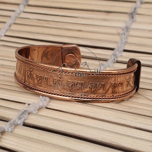 May include: A copper bracelet with a magnetic clasp. The bracelet is engraved with a Sanskrit mantra in a circular pattern.
