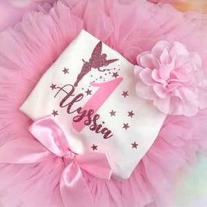 May include: A pink tutu with a white top featuring a glittery fairy design, the number 1, and the name "Alyssia". The outfit includes a large pink bow and a pink flower. The background is a soft pink.