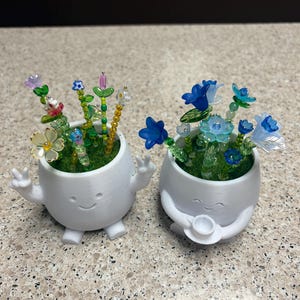 May include: Two white 3D-printed planters with smiling faces and arms. The left planter has arms raised in a peace sign and holds colorful beaded flowers. The right planter holds a small cup and features blue beaded flowers. Both have green moss.