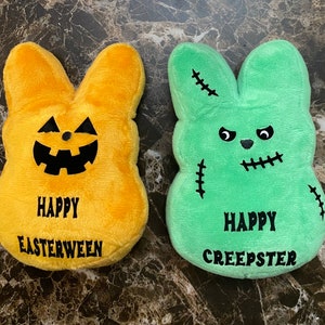 May include: Two plush Easter bunny shaped pillows. One is yellow with a jack-o-lantern face and the text "Happy Easterween". The other is green with a stitched-up monster face and the text "Happy Creepster".