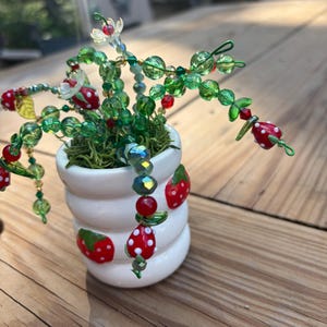 May include: A white ceramic pot with three horizontal ridges and painted red strawberry motifs, filled with green moss. Emerging from the moss are wire stems adorned with green glass beads, small clear flower beads, and red strawberry-shaped beads.
