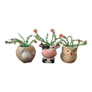 May include: Three small ceramic planters shaped like a sheep, a cow, and an owl. Each planter contains a decorative plant made of green beads and small pink flowers. The sheep is beige, the cow is black and white with a pink snout, and the owl is brown.