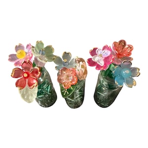 May include: Three small, clear glass jars filled with green beads, each topped with a cluster of translucent, multi-colored acrylic flowers. The flowers feature gold-toned edges and centers, showcasing shades of pink, orange, blue, and clear.