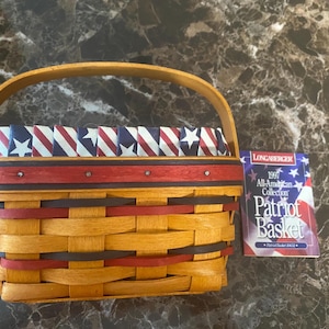 May include: A woven basket with a wooden handle, featuring red, white, and blue accents. The basket has a patriotic theme, with a patterned fabric lining the top edge. A Longaberger tag is visible.