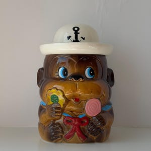 Pode incluir: Um pote de biscoitos de cerâmica vintage em forma de macaco usando um chapéu de marinheiro com um emblema de âncora. O macaco tem olhos azuis, segura um pirulito e um doce em forma de flor, e é adornado com um laço vermelho e detalhes em azul. O pote é marrom e creme.