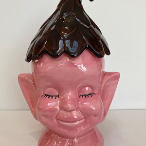 Könnte beinhalten: Eine rosafarbene Keramik-Keksdose in Form eines lächelnden Elfen mit geschlossenen Augen. Der Elf hat große Ohren und einen braunen Hut mit einer spitzen Spitze.