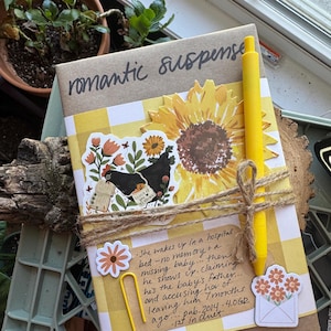 May include: A book wrapped with twine, featuring the words "romantic suspense". It's decorated with a sunflower, stickers, and a yellow pen. The book has a yellow and white checkered pattern. Text on the book reads: "She wakes up in a hospital bed..."