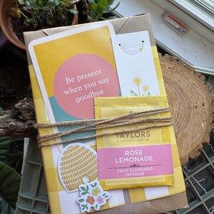 May include: A gift package wrapped in brown paper and yellow gingham. It features a card with the text "Be present when you say goodbye", a tea bag, and decorative stickers. The package is tied with twine.