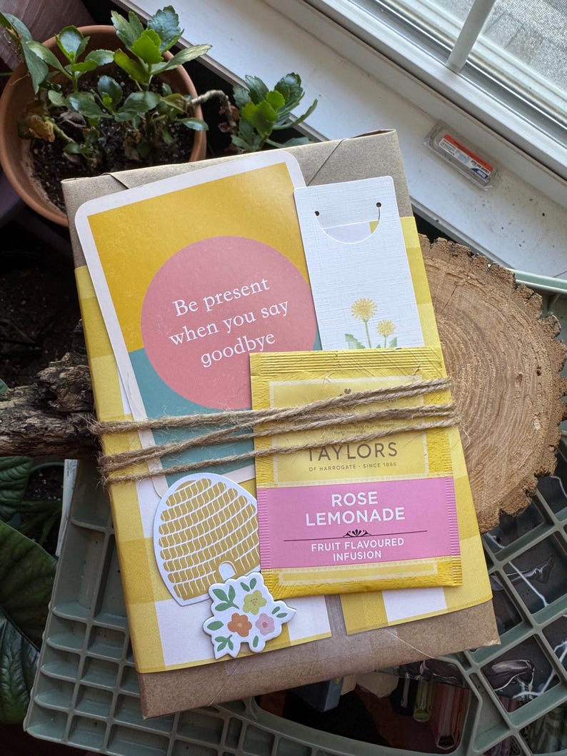 May include: A gift package wrapped in brown paper and yellow gingham. It features a card with the text "Be present when you say goodbye", a tea bag, and decorative stickers. The package is tied with twine.