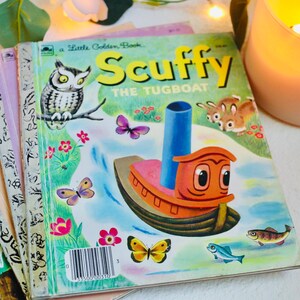 May include: A collection of vintage children's books, including "Scuffy the Tugboat." The cover of "Scuffy" shows an orange tugboat, an owl, butterflies, and the title in yellow. Other books have colourful covers. A lit candle and greenery are in the background.