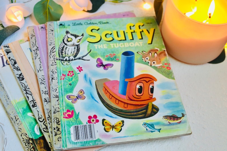 May include: A collection of vintage children's books, including "Scuffy the Tugboat." The cover of "Scuffy" shows an orange tugboat, an owl, butterflies, and the title in yellow. Other books have colourful covers. A lit candle and greenery are in the background.