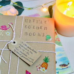 May include: A parcel of "Little Golden Books" wrapped in brown paper and tied with white string. The tag reads "5 Little Golden Books Published in the 50s, 70s & 80s. Fair condition - loved by several generations!" Decorative stickers of a flamingo, cactus, and pineapple are on the package. A lit candle is in the background.