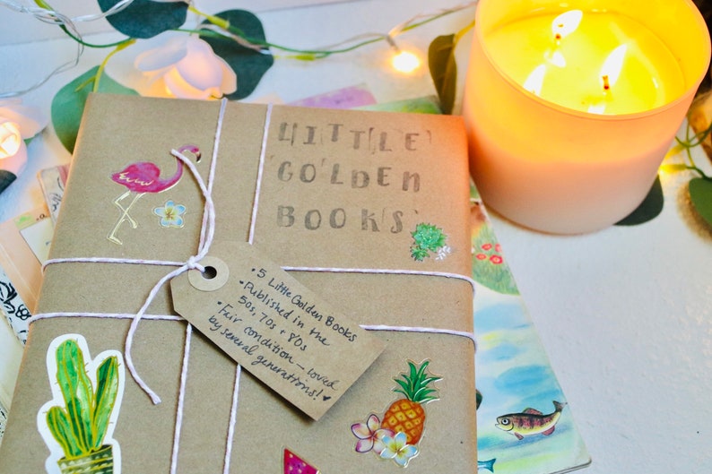 May include: A parcel of "Little Golden Books" wrapped in brown paper and tied with white string. The tag reads "5 Little Golden Books Published in the 50s, 70s & 80s. Fair condition - loved by several generations!" Decorative stickers of a flamingo, cactus, and pineapple are on the package. A lit candle is in the background.