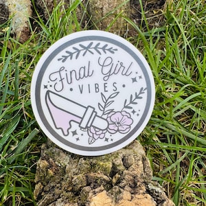 May include: A round sticker with the text "Final Girl Vibes" in a decorative font. The sticker features a knife with pink dripping accents, floral elements, and stars. The design is set against a white background with a gray border.