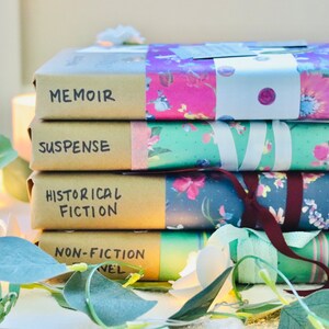 May include: Four books wrapped in brown paper and floral patterned paper. The books are labelled "Memoir", "Suspense", "Historical Fiction", and "Non-Fiction Novel". Fairy lights and greenery are arranged around the books.