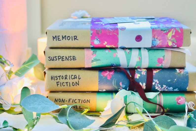 May include: Four books wrapped in brown paper and floral patterned paper. The books are labelled "Memoir", "Suspense", "Historical Fiction", and "Non-Fiction Novel". Fairy lights and greenery are arranged around the books.