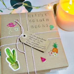 May include: A bundle of Little Golden Books wrapped in brown paper, decorated with stickers of a flamingo, cactus, and pineapple. The books are tied with string and a tag that reads "5 Little Golden Books published in the 50s, 70s & 80s". A lit candle is in the background.