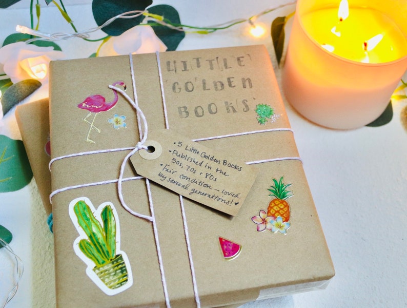 May include: A bundle of Little Golden Books wrapped in brown paper, decorated with stickers of a flamingo, cactus, and pineapple. The books are tied with string and a tag that reads "5 Little Golden Books published in the 50s, 70s & 80s". A lit candle is in the background.