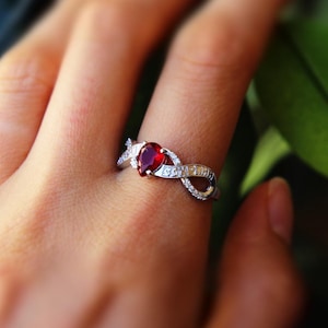 May include: A silver-toned ring featuring a pear-shaped red gemstone at the centre, flanked by an infinity-shaped band design encrusted with small, clear, sparkling stones along the metal surface, creating a continuous loop effect.