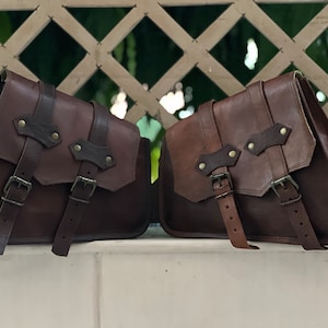 May include: Two brown leather motorcycle saddlebags with buckles and straps. The bags are made of a dark brown leather with a lighter brown leather trim.