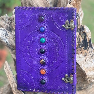 May include: Purple leather journal with a decorative embossed design and a clasp closure. Seven colorful gemstones are inlaid along the spine.
