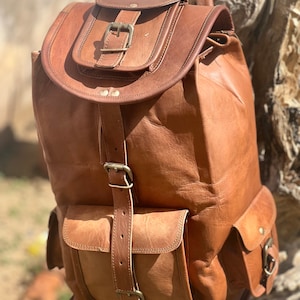 Może przedstawiać: Brązowy skórzany plecak z wieloma kieszeniami i klamrami. Plecak ma klapę z zapięciem na klamrę i dużą kieszeń z przodu.