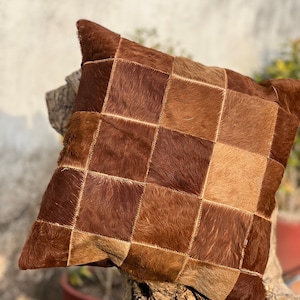 Puede incluir: Una almohada de cuero patchwork marrón y beige con un patrón cuadrado. La almohada está hecha de piel de vaca y tiene una textura suave y peluda.