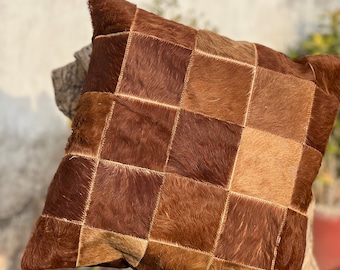 May include: A brown and tan patchwork leather pillow with a square pattern. The pillow is made of cowhide and has a soft, fuzzy texture.