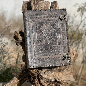 May include: A brown leather journal with an embossed tree of life design and two metal clasps. The journal is sitting on a piece of driftwood.