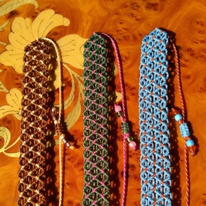 Puede incluir: Tres pulseras de macramé hechas a mano con cordones de diferentes colores. Las pulseras son marrón y blanco, verde y rosa, y azul y marrón. Las pulseras están atadas con un nudo al final y tienen cuentas.