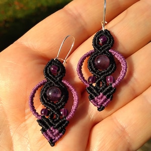 May include: A pair of black and purple macrame earrings with amethyst stones. The earrings are made using a knotting technique and feature a teardrop shape.