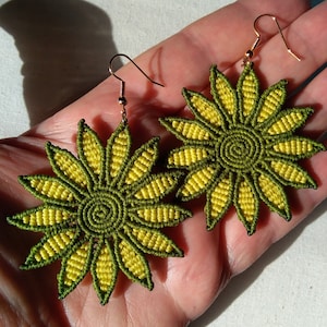 Op de afbeelding: Een paar groene en gele zonnebloem oorbellen. De oorbellen zijn gemaakt van macramé en hebben een gouden haak.