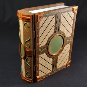 Op de afbeelding: Een decoratieve doos in de vorm van een boek met een houten deksel, groene en gouden accenten en een ronde uitsparing aan de voorkant. De doos is gesloten en lijkt van hout te zijn.